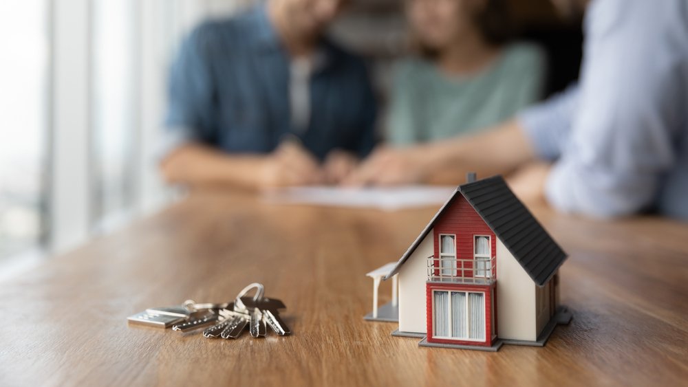 Peniaze na vlastné bývanie - ilustračný obrázok, kľúč a malý domček na stole, v pozadí podpisovanie hypotekárnej zmluvy
